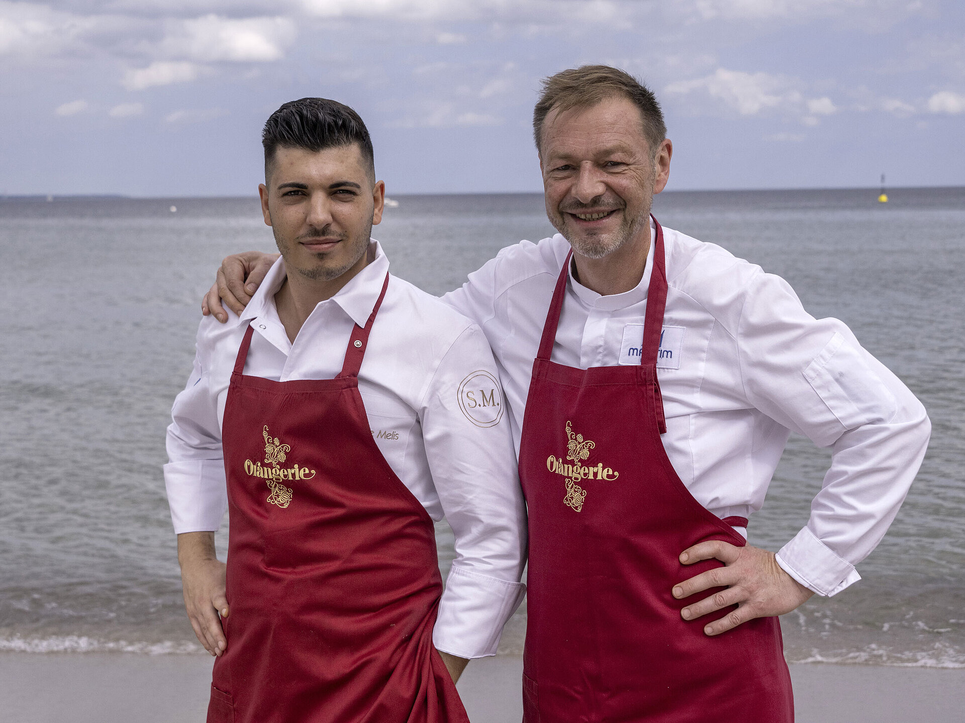 Küchenchefs Simone Melis und Thomas Lemke mit roten Schürzen am Meer vor dem Maritim Hotel Timmendorfer Strand.